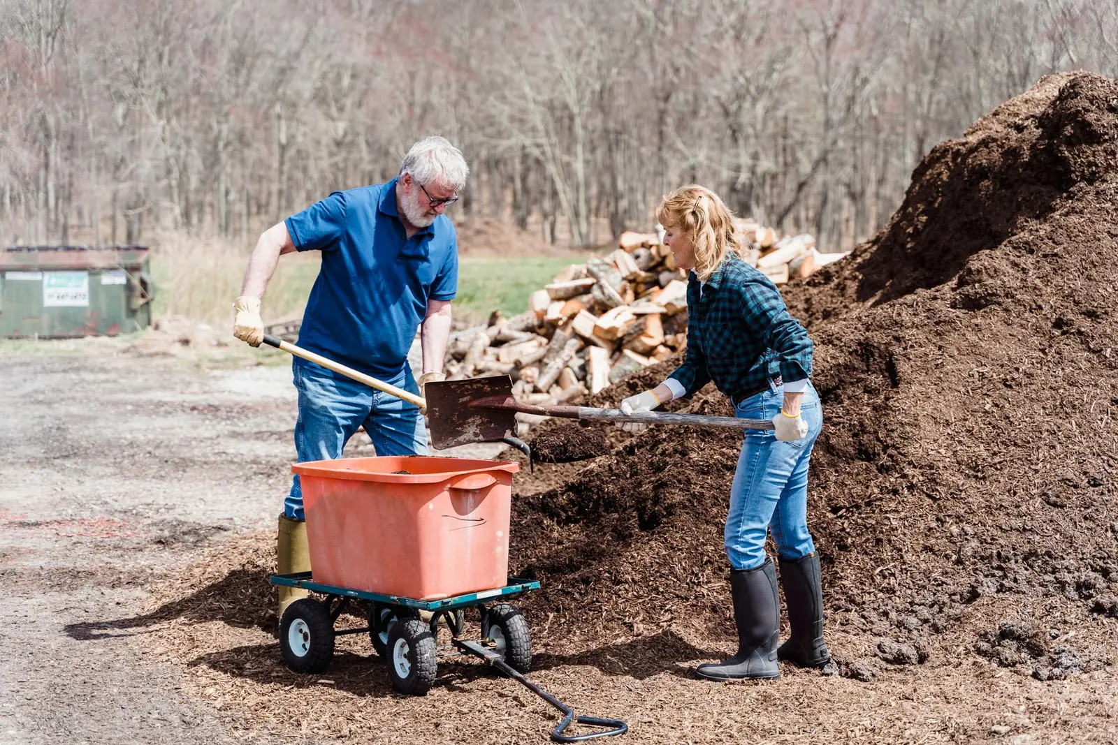 Two gardeners shoveling loose brown material into an orange garden cart beside large piles