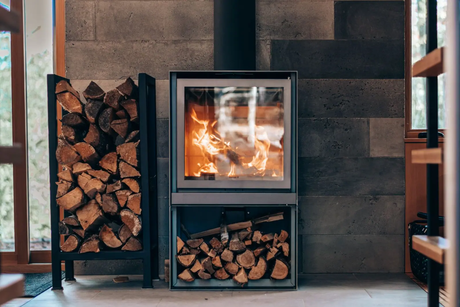 A black glass-fronted stove burning logs beside stacked firewood